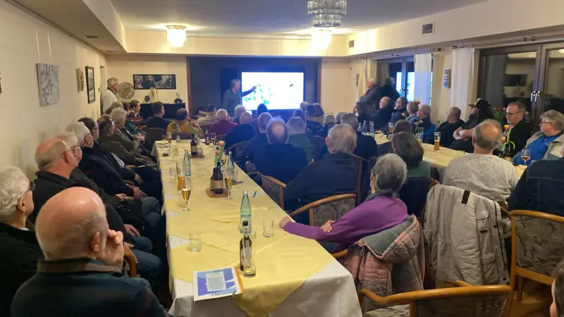 Raum von hinten fotografiert, im Hintergrund steht der Vortragende vor einem Bildschirm, im Vodergrund sitzen die Zuhörerinnen und Zuhörer an Tischen